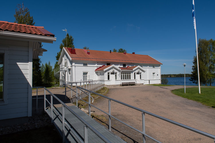 Kuvassa valkoinen vanha leirikeskus rakennus järven rannalla. lipputangossa on suomenlippu.